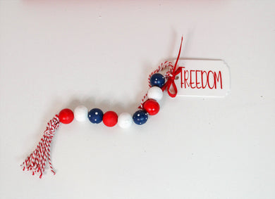 Red, white and blue wood bead garland with tassel and tag, which features the word freedom