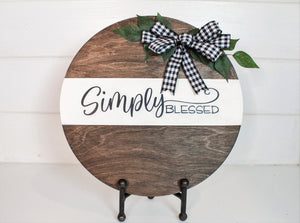 Round wood 12 inch sign displays the saying, "Simply Blessed." Sign is stained with an espresso finish and features a buffalo check bow and greenery
