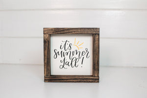 Wood framed sign featuring the saying, "It's summer y'all." Frame is stained espresso and sign is a cream background with black lettering and a yellow sun accent