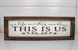 Wood framed sign with the saying, "Our life, our story our home. This is us." Frame is stained with an espresso finish around a cream background and black lettering and accents